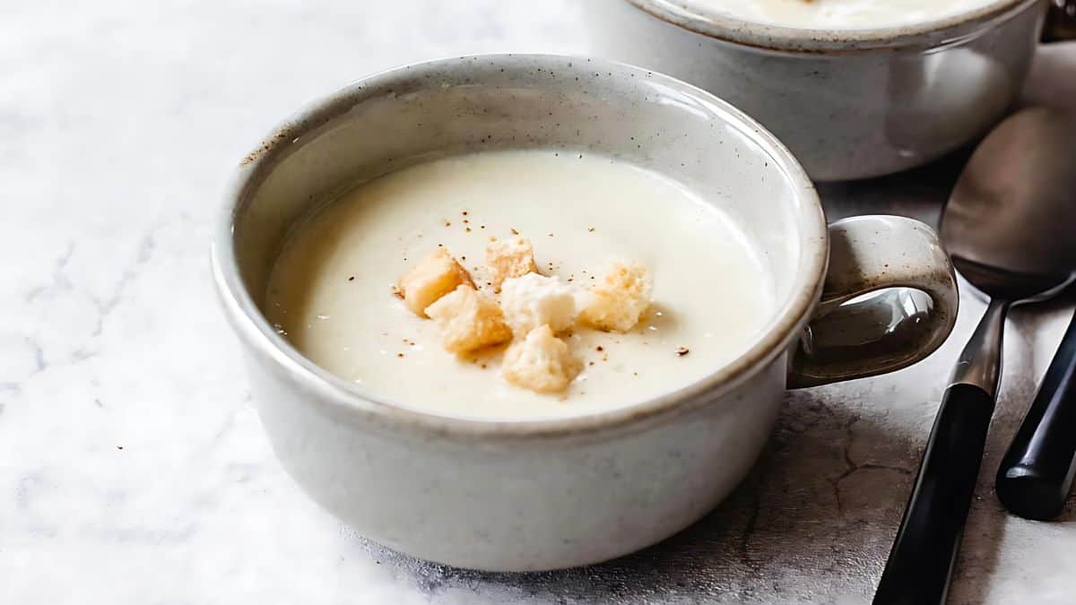 A ceramic bowl filled with creamy soup, garnished with croutons and a sprinkle of seasoning. The bowl is set on a light-colored surface, and a spoon rests next to it. Another identical bowl is partially visible in the background.