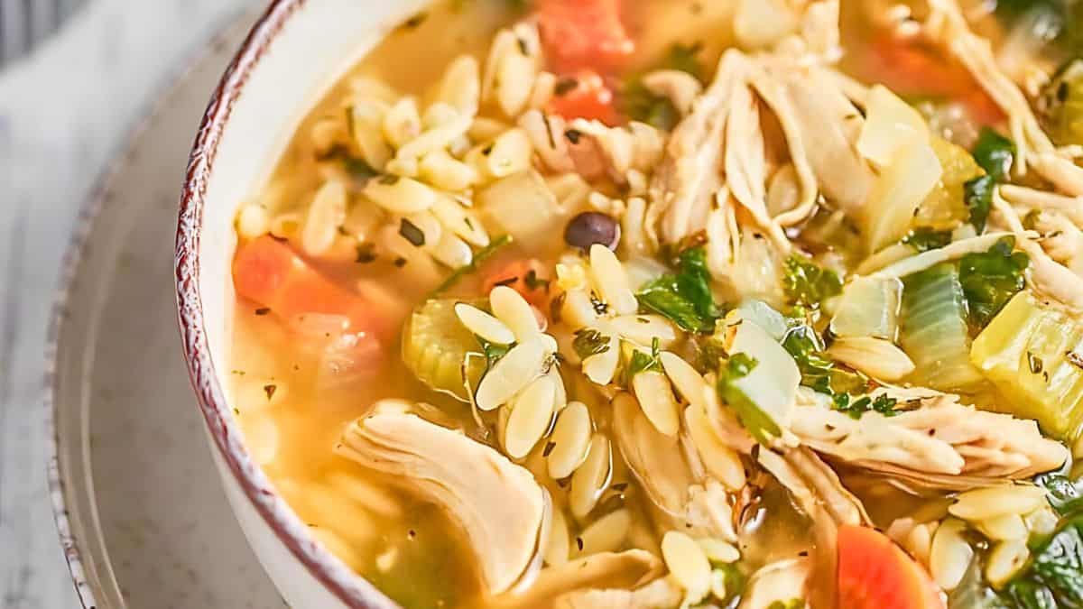 A bowl of chicken orzo soup filled with shredded chicken, orzo pasta, chopped carrots, celery, and herbs in a clear broth. The soup is served in a white ceramic bowl on a light surface.