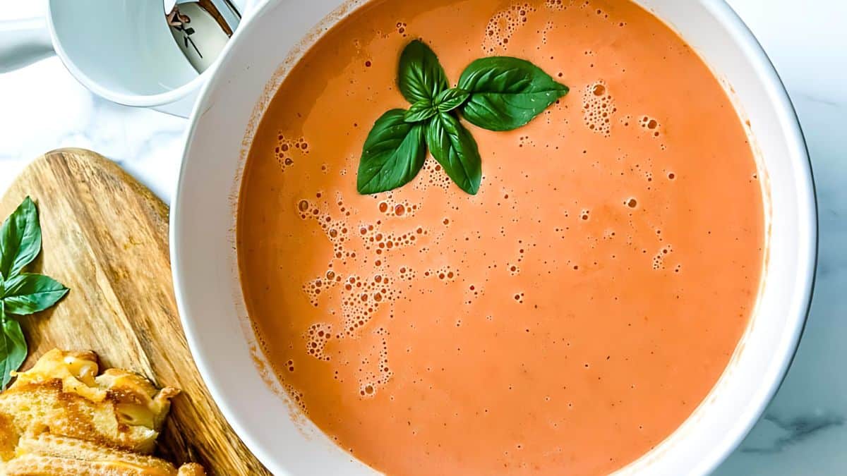 A bowl of creamy tomato soup garnished with three fresh basil leaves at the center. To the side, on a wooden board, are two halves of a grilled cheese sandwich with melted cheese oozing out. A white teacup can be seen in the background.