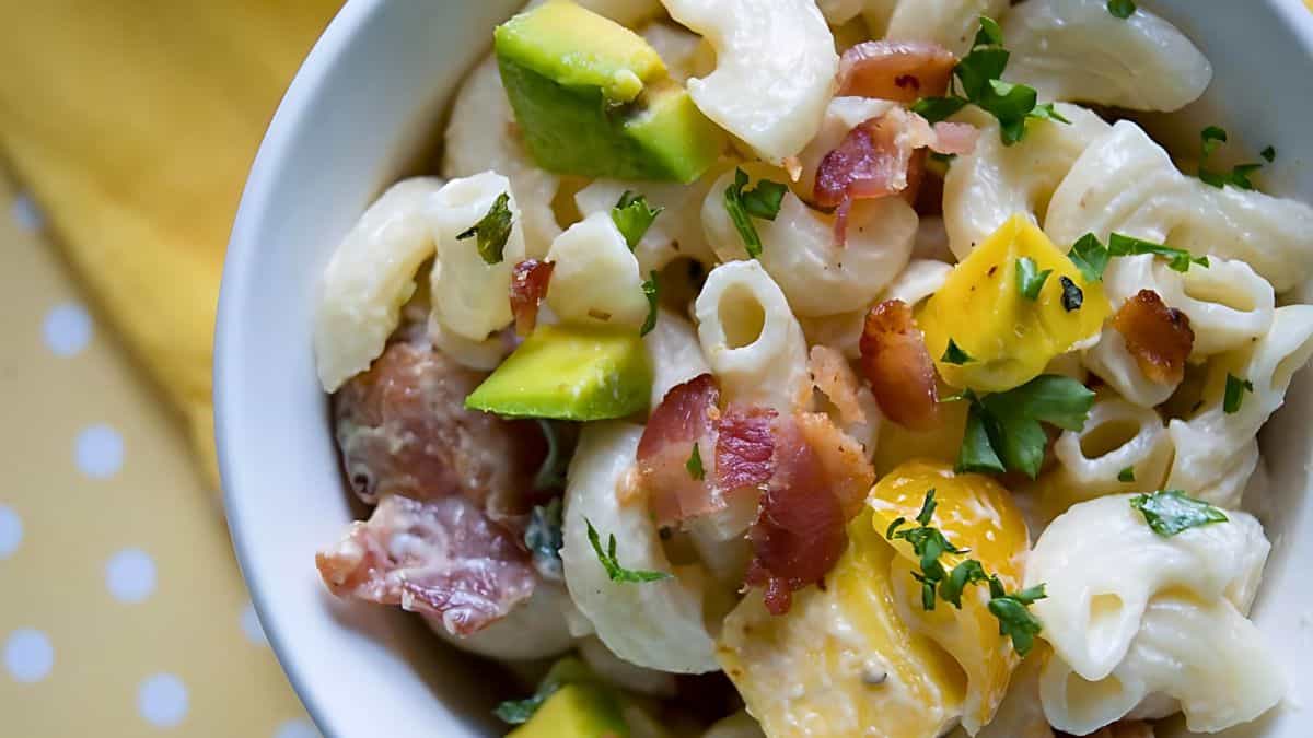 A white bowl filled with creamy pasta salad. The pasta is mixed with avocado chunks, cooked bacon pieces, and garnished with chopped parsley. The bowl is placed on a yellow and white polka dot tablecloth.