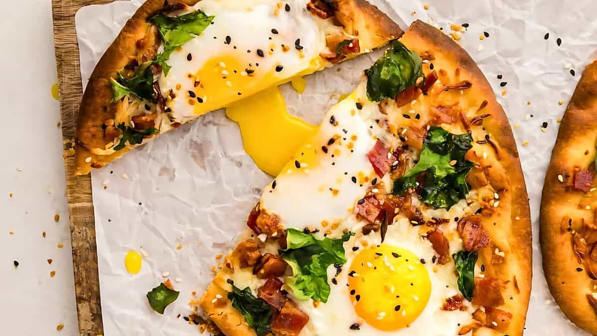 A delicious breakfast pizza with a golden, runny egg yolk, leafy greens, crispy bacon, and sprinkled sesame seeds on a thin, crispy crust placed on parchment paper. One slice has been cut and slightly pulled away, showing the tempting melted cheese and toppings.