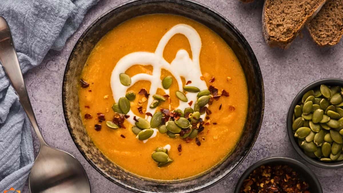 A bowl of creamy orange soup garnished with a swirl of white cream, pumpkin seeds, and red chili flakes. Surrounding the bowl are slices of crusty bread, a spoon on a napkin, and two small bowls, one containing pumpkin seeds and the other filled with chili flakes.