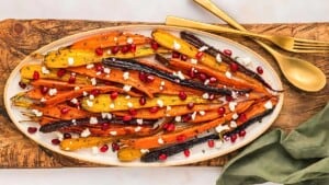 A white oval platter holds a colorful dish of roasted rainbow carrots garnished with crumbled feta cheese and pomegranate seeds. The platter rests on a wooden board along with gold-colored cutlery. A green cloth napkin is placed to the side.