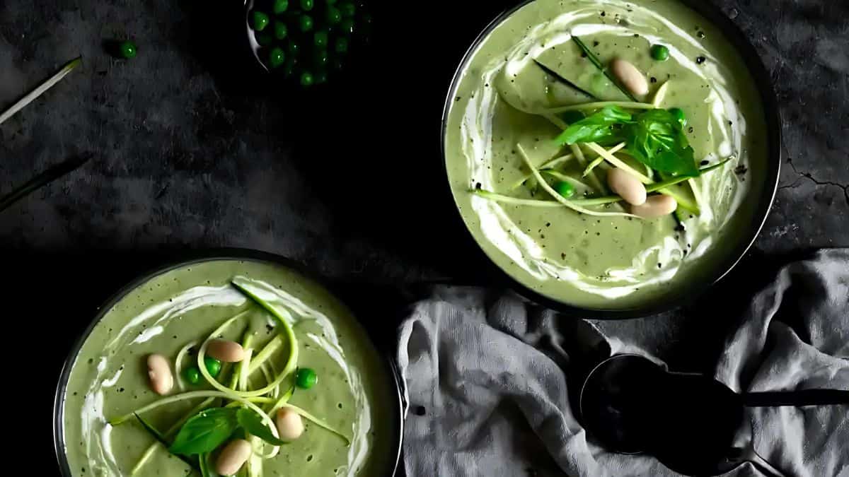 Two bowls of creamy green soup garnished with white beans, peas, and fresh greens sit on a dark surface. A grey cloth and a black spoon are next to the bowls, adding to the rustic presentation. The overall ambiance is elegant and moody.