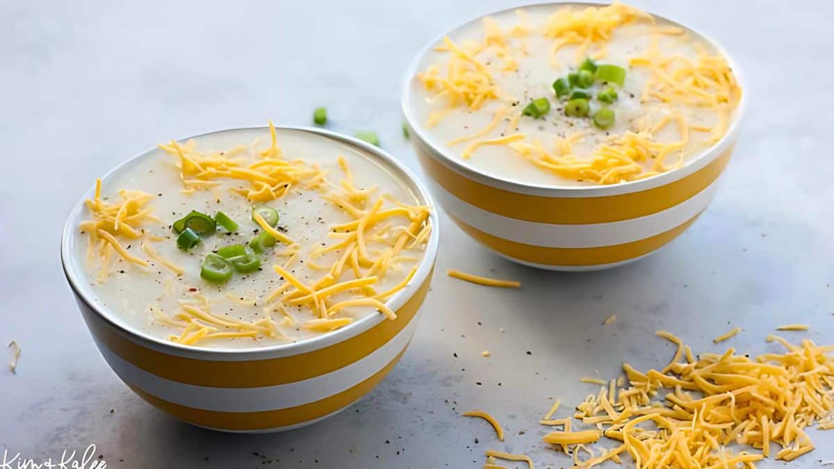 Two bowls of creamy soup topped with shredded cheese and chopped green onions, placed on a light surface. A small pile of cheese is scattered nearby. The bowls have yellow and white stripes.