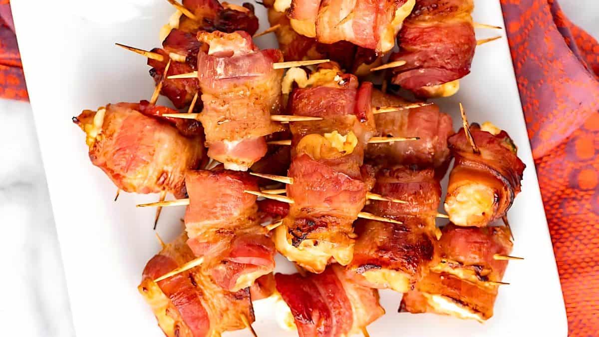 A close-up view of a white rectangular plate filled with bacon-wrapped appetizers. The appetizers are secured with toothpicks and placed attractively on the plate. A red patterned napkin is partially visible on the side.