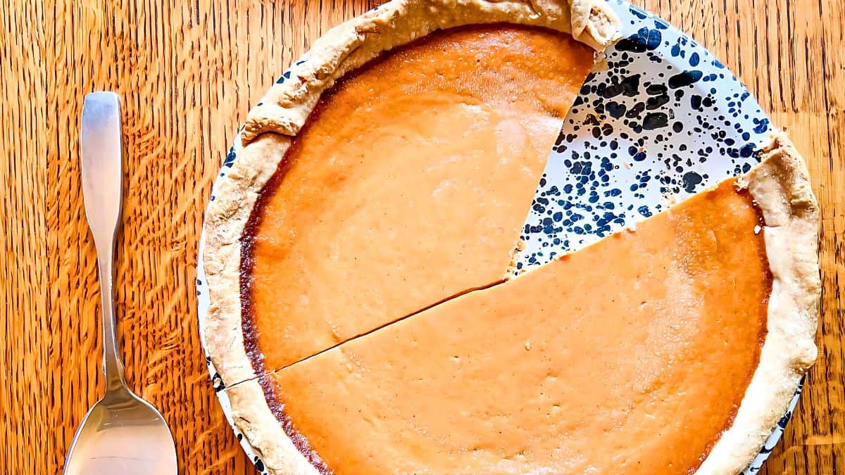 A top view of a partially sliced pumpkin pie on a white and blue speckled ceramic plate, placed on a wooden table. A serving utensil lies to the left of the plate. The pie has a golden-brown crust and smooth, creamy filling.
