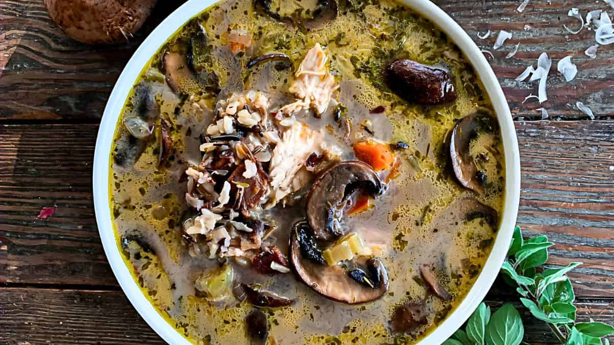 A bowl of homemade chicken and wild rice soup, garnished with mushroom slices, carrots, celery, and fresh herbs. The soup has a golden broth and is presented on a rustic wooden table with fresh mushrooms and herbs beside it.