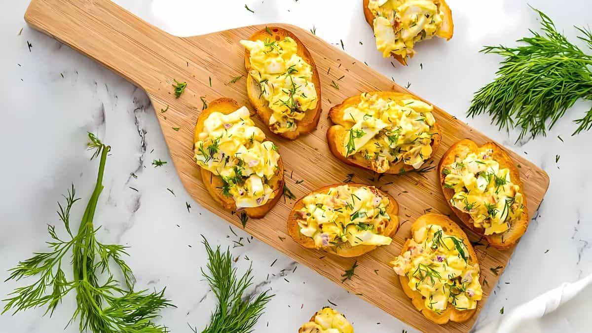 A wooden cutting board with six pieces of toasted bread topped with a creamy egg salad garnished with fresh dill. Fresh dill sprigs are scattered on the marble countertop around the cutting board, adding a touch of greenery to the presentation.