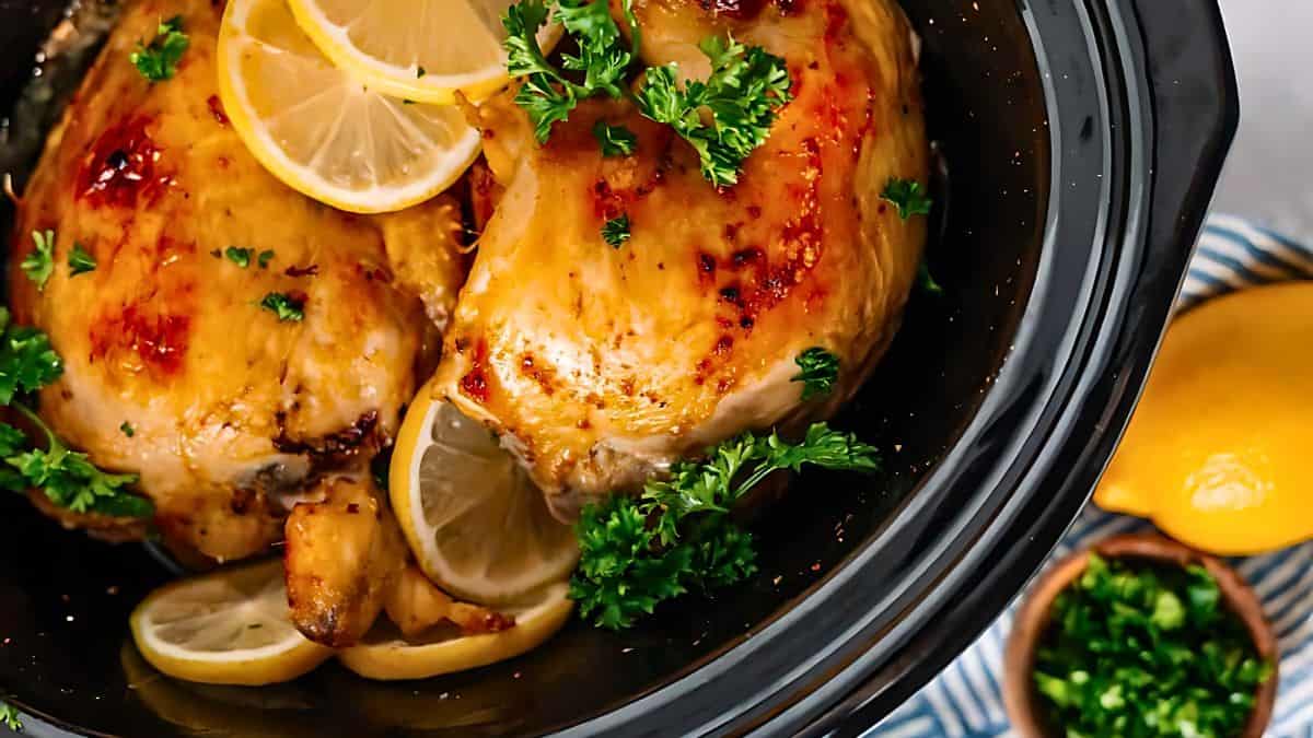 Close-up of roasted chicken in a black pot, garnished with lemon slices and fresh parsley. Brightly lit, the dish sits on a striped cloth, with a bowl of chopped herbs and a lemon nearby.