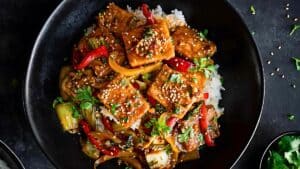 A colorful dish of crispy tofu topped with sesame seeds, mixed with red and yellow bell peppers, bok choy, and cilantro, all served over white rice in a black bowl.