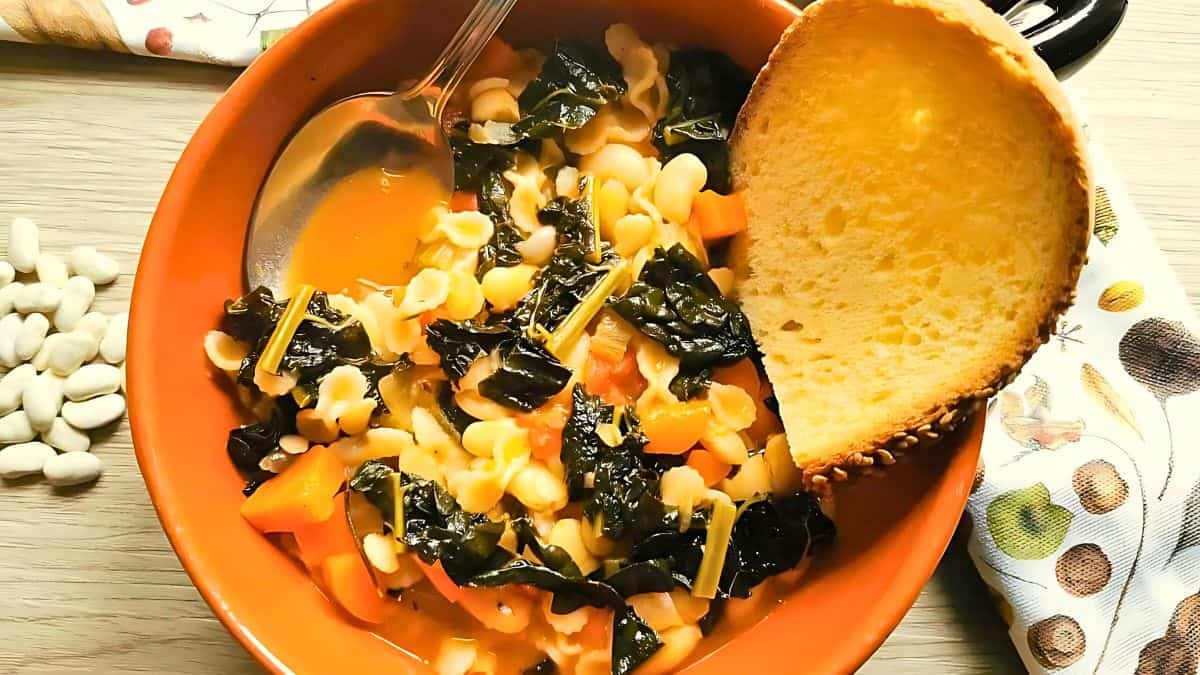 A bowl of vegetable and bean soup containing kale, carrots, pasta, and white beans, garnished with a large slice of crusty bread. A spoon rests inside the bowl on a wooden surface, next to a patterned cloth and scattered white beans.