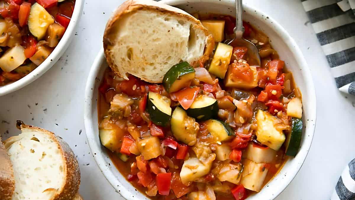 A bowl of ratatouille is topped with a slice of crusty bread. The dish is filled with a colorful mix of vegetables, including zucchini, tomatoes, red bell peppers, and onions. In the background, partially visible are another bowl of ratatouille and another slice of bread.