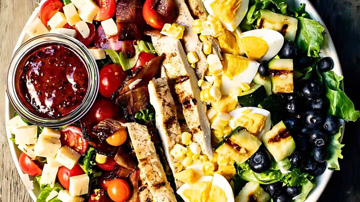 A fresh, vibrant salad in a bowl featuring sliced grilled chicken, boiled eggs, cherry tomatoes, blueberries, corn kernels, diced cheese, grilled zucchini, lettuce, and a jar of red dressing on the side. The ingredients are neatly arranged.