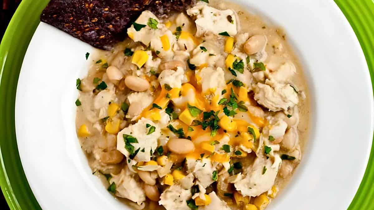 A bowl of creamy white chicken chili with chunks of chicken, white beans, corn, and shredded cheese, garnished with chopped parsley. A piece of dark bread rests on the side, all set on a green plate.