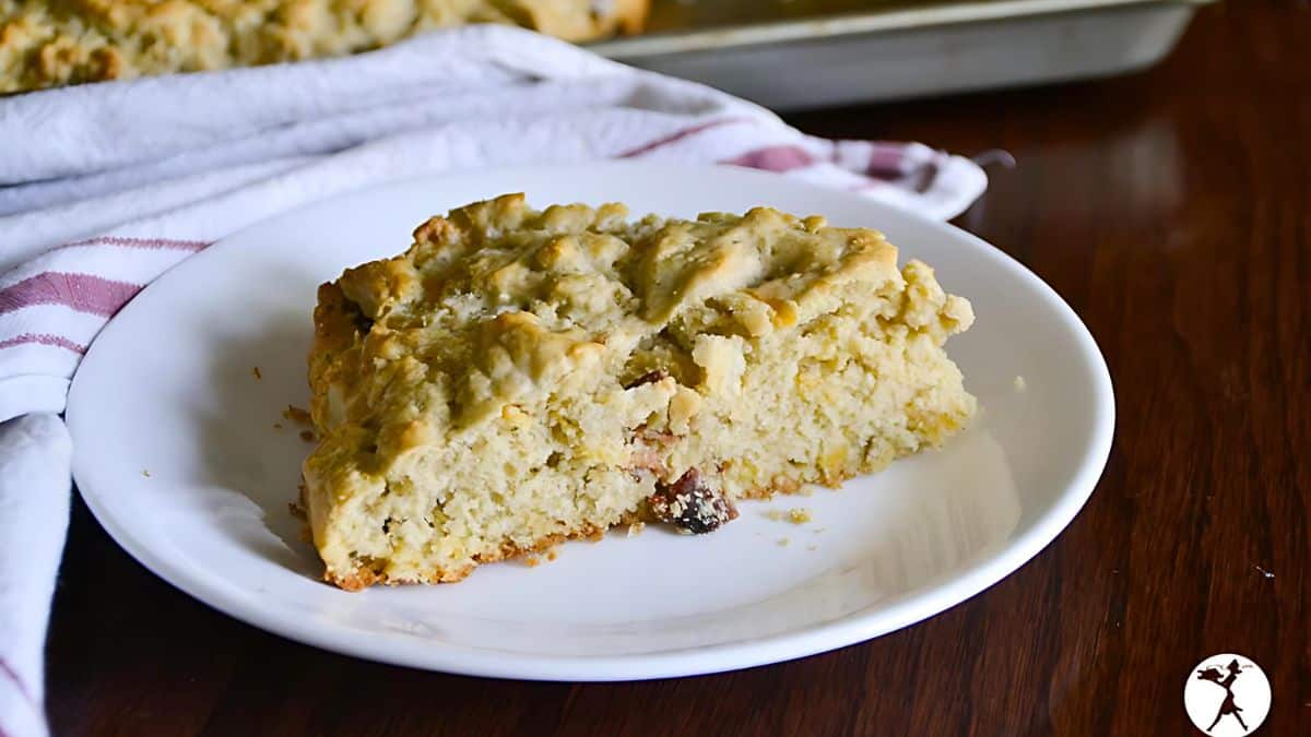 A slice of crumbly, golden-brown scone sits on a white plate, placed on a wooden table. A portion of a striped cloth is visible beneath the plate. The scone appears to have pieces of dried fruit, possibly raisins or cranberries.