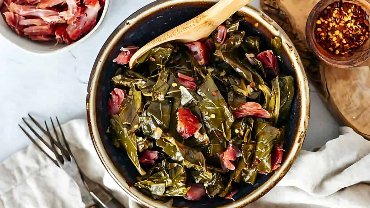 A bowl of cooked collard greens mixed with pieces of ham, garnished with red pepper flakes. A wooden spoon is resting in the bowl. Nearby, there is a small bowl of additional ham and a wooden dish with more red pepper flakes.