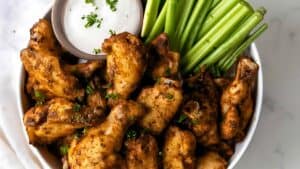 A bowl filled with seasoned chicken wings garnished with parsley, accompanied by a side of celery sticks and a small bowl of dipping sauce.