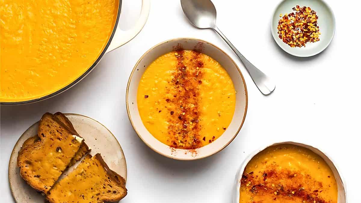 A bowl of creamy orange soup sprinkled with red pepper flakes is the focal point. Surrounding the main bowl are a pot of the same soup, a spoon, a smaller bowl with red pepper flakes, and a plate with two slices of toasted bread. All items are on a white surface.