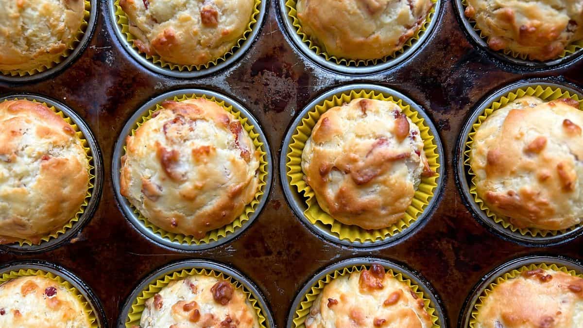 A close-up view of freshly baked muffins in a dark muffin tray. Each muffin has a golden-brown top and is placed in a yellow paper liner. The muffins look fluffy and delicious, with some having visible pieces of ingredients mixed in.