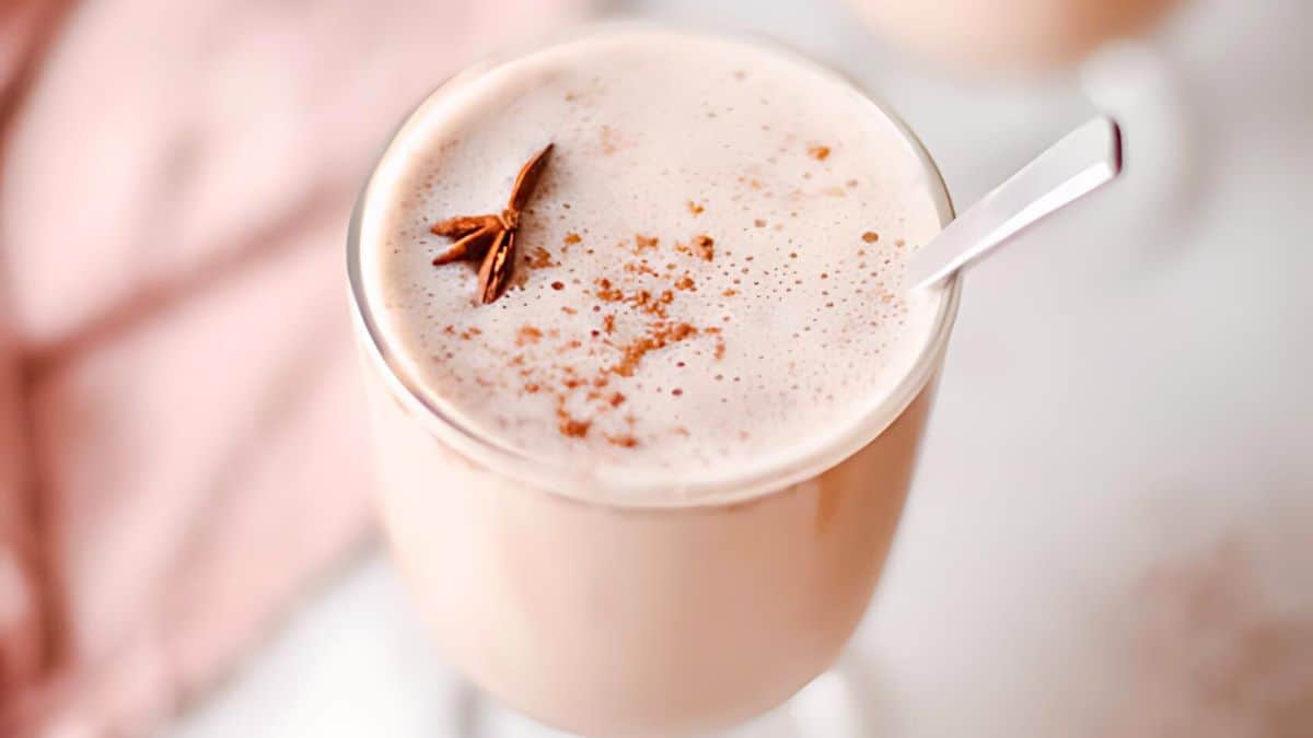 A creamy chai latte in a glass, garnished with a sprinkle of cinnamon and a star anise, with a spoon resting on the side. A soft pink cloth is blurred in the background.