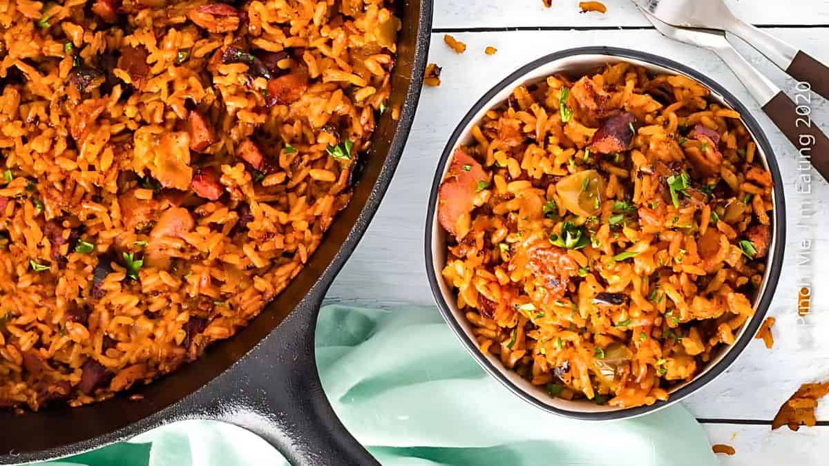 A skillet filled with a savory rice dish featuring sausage and vegetables is shown on the left, while a bowl containing the same rice dish is placed on the right. Both are set on a white wooden surface with a light green cloth nearby and a fork and spoon beside the bowl.