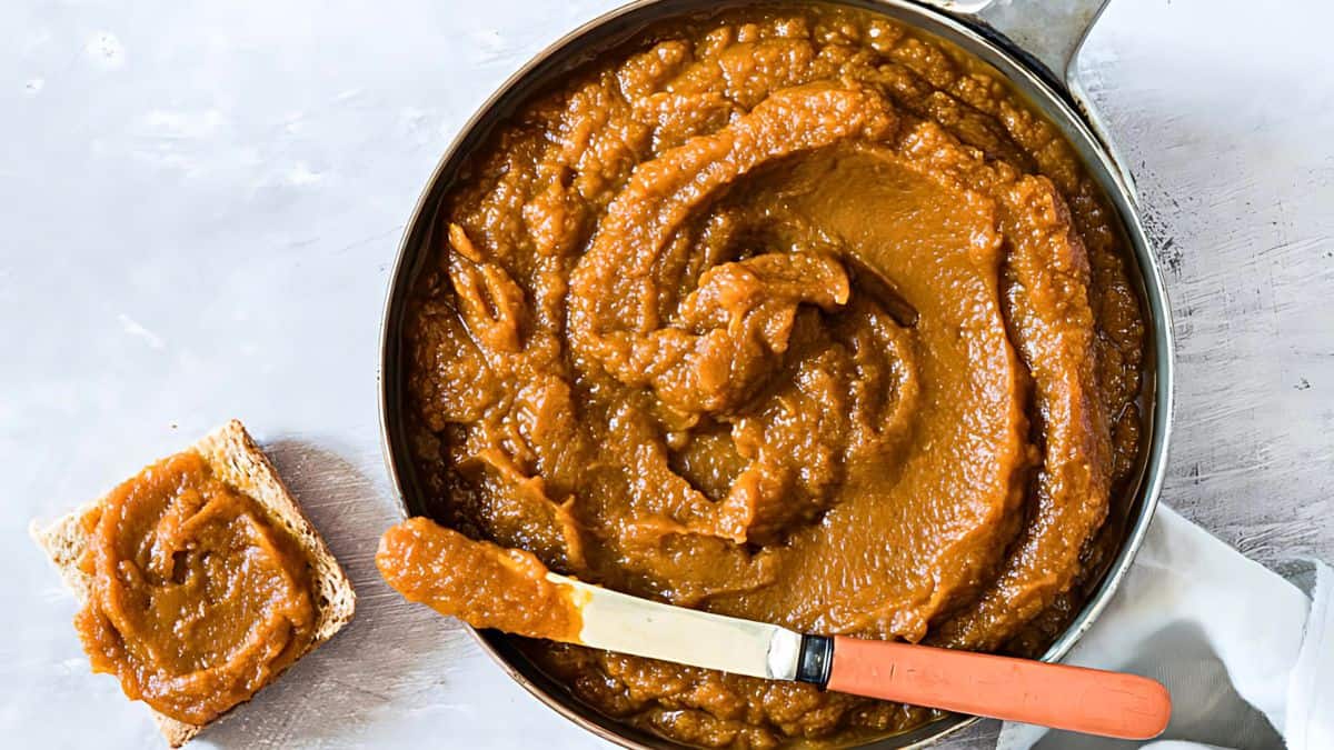 A pot filled with thick, smooth apple butter sits on a light surface. A knife with a wooden handle, coated with apple butter, rests partly on the pot. A slice of bread spread with apple butter is next to the pot.
