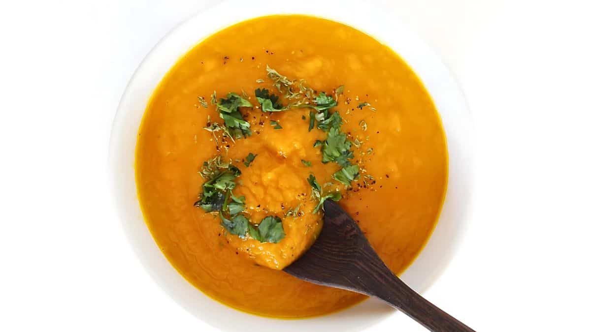 A white bowl filled with bright orange carrot soup, garnished with chopped green cilantro and black pepper. A dark wooden spoon partially submerged in the soup is visible at the bottom right corner. The background is a plain white surface.
