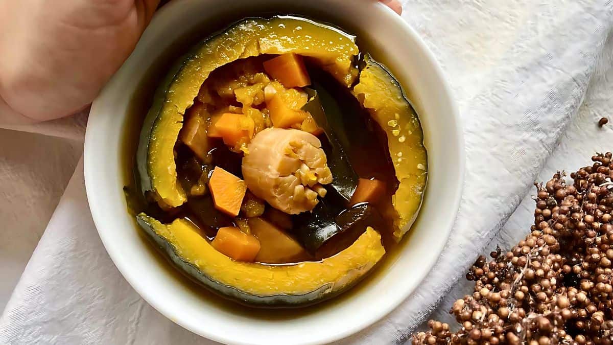 A hand holding a white bowl of savory stew. The dish contains slices of pumpkin, chunks of carrot, a scallop, and other ingredients in a clear broth. The bowl is on a light-colored cloth next to a cluster of small, grain-like seeds.