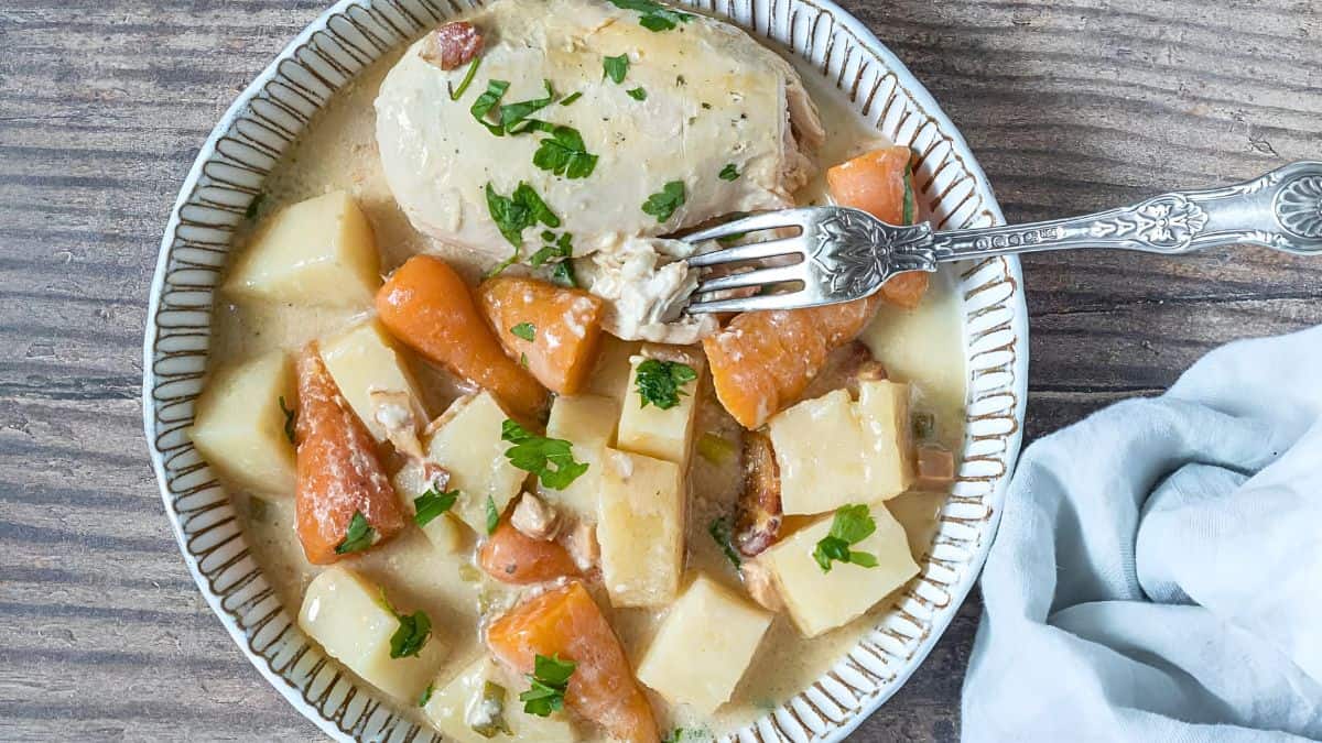 A plate of creamy chicken stew with diced carrots and potatoes, garnished with chopped parsley. A fork rests on the plate. A white napkin is partially visible to the right on a wooden table surface.