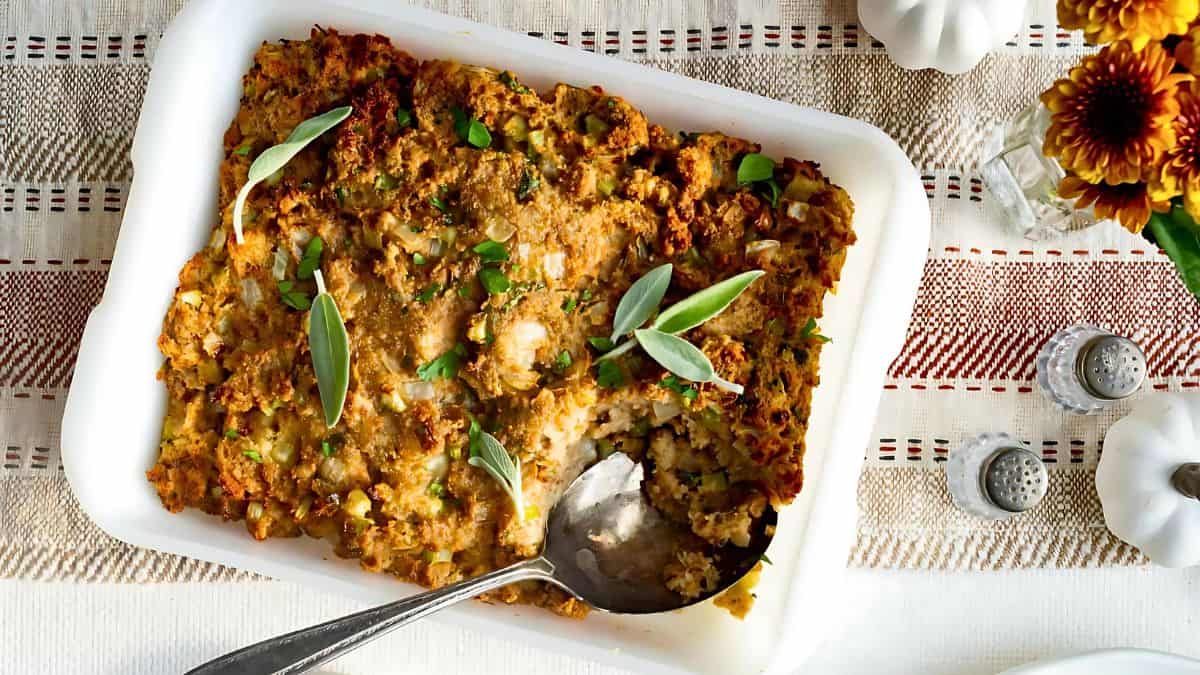 A rectangular dish of baked stuffing garnished with fresh herbs is placed on a patterned tablecloth. A serving spoon is resting inside the dish, indicating a portion has been taken. The table is decorated with flowers and salt and pepper shakers.