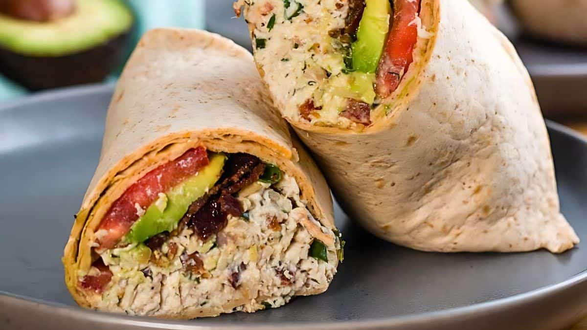 Close-up of a sliced chicken wrap with fresh ingredients. The wrap includes chunks of chicken, avocado slices, tomatoes, and lettuce, all encased in a tortilla. The texture and colors showcase the freshness of the ingredients.