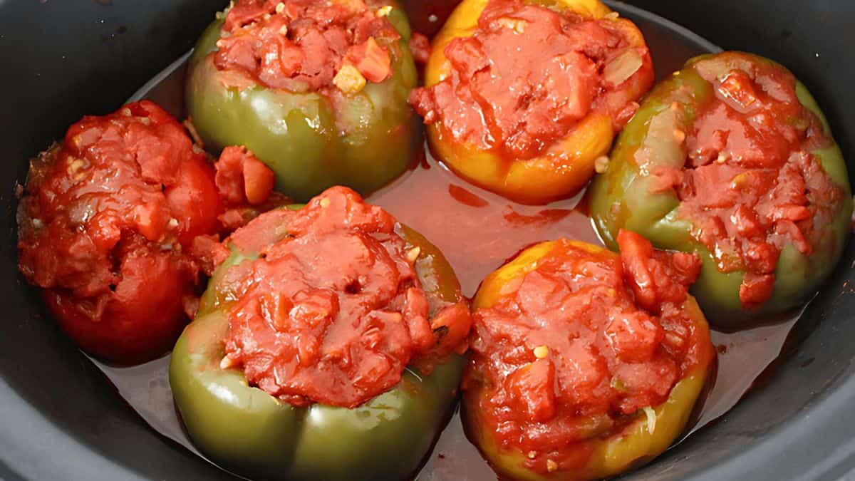 Six colorful bell peppers filled with a tomato-based mixture are placed in a black crockpot. The peppers are red, yellow, and green, and the mixture inside appears chunky and rich in tomato sauce.