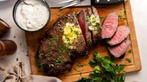 Sliced steak on a cutting board with butter and chopped herbs on top, accompanied by a bowl of creamy sauce. Fresh parsley and wooden salt and pepper shakers are nearby.