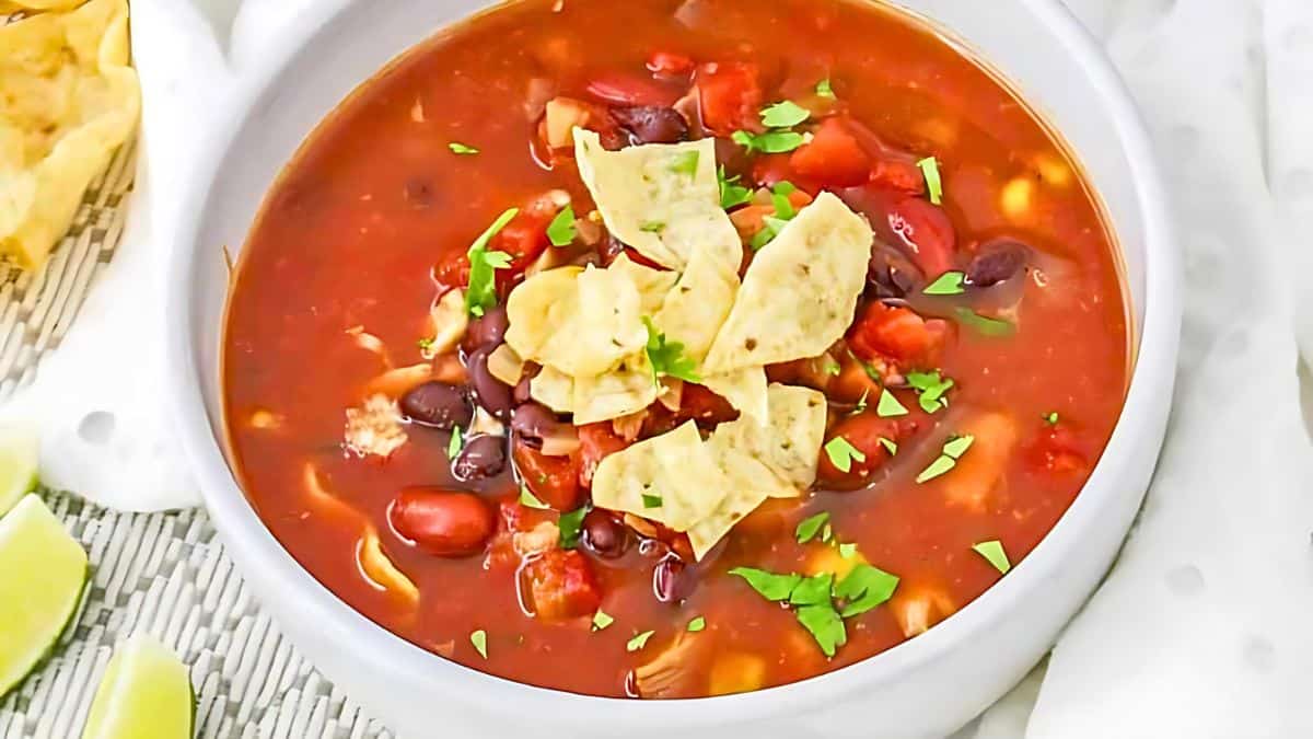 A bowl of red tortilla soup garnished with tortilla chips and chopped cilantro. The soup contains black beans, kidney beans, corn, and visible chunks of tomato. Lime wedges and chips are to the side on a woven mat.