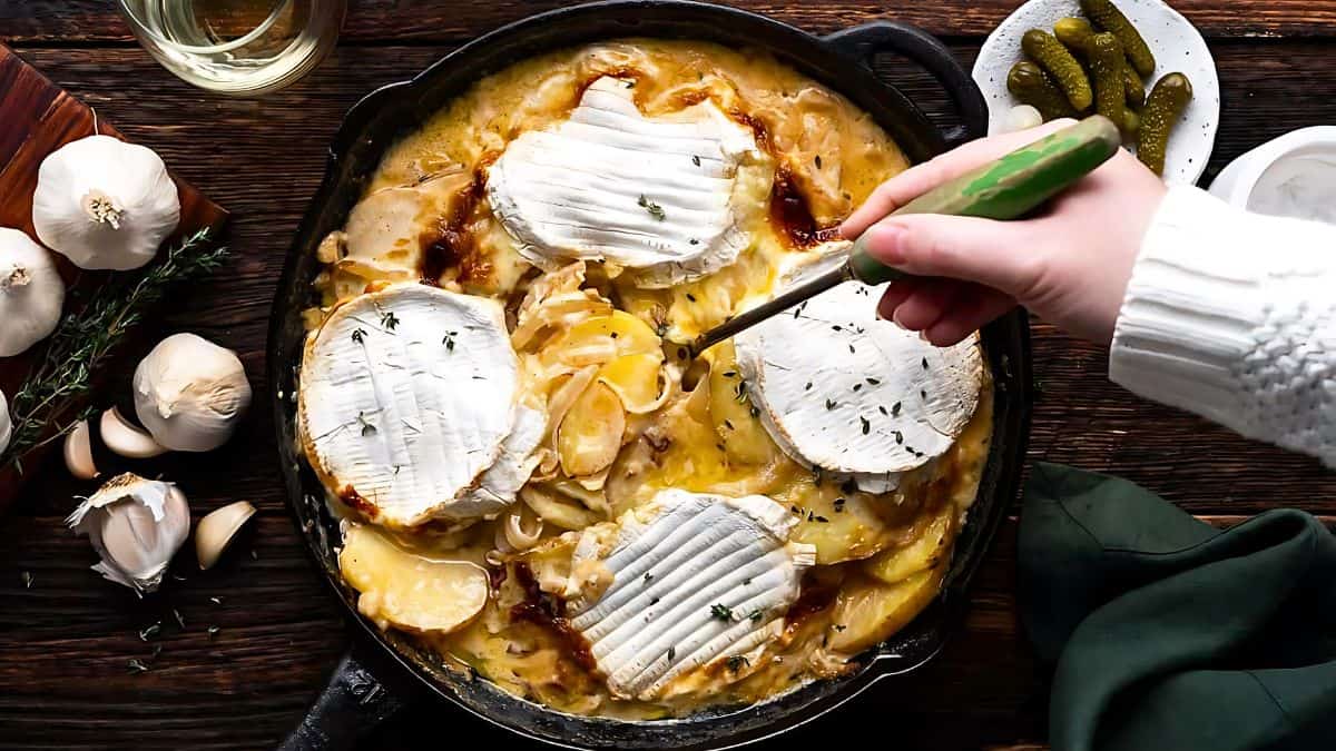 A skillet filled with creamy potato gratin is topped with rounds of melting cheese. A hand holds a green spoon, scooping some of the dish. Surrounding the skillet are cloves of garlic, sprigs of herbs, pickles on a plate, and a glass of white wine.