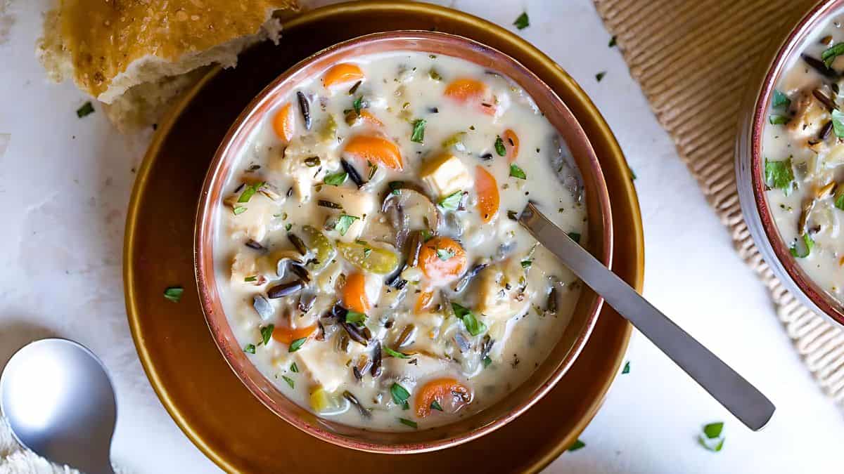 A bowl of creamy soup filled with wild rice, chopped carrots, mushrooms, and chunks of chicken, garnished with herbs. A spoon rests in the bowl, and a piece of bread is placed nearby on the table.