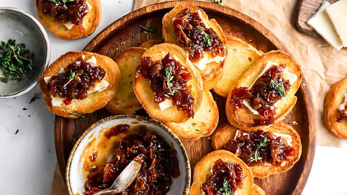 A wooden tray filled with toasted baguette slices topped with creamy cheese, caramelized onion jam, and garnished with fresh herbs. A bowl of the caramelized onion jam with a spoon is placed next to the tray, along with a sprig of herbs and cheese slices.
