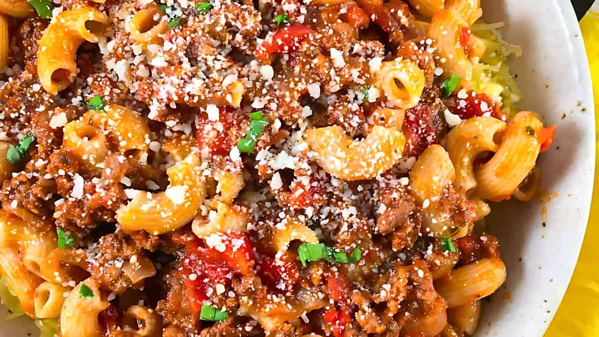 A bowl of pasta topped with a rich meat sauce, diced tomatoes, and a sprinkle of grated cheese. The pasta is garnished with chopped green herbs for added color and flavor.