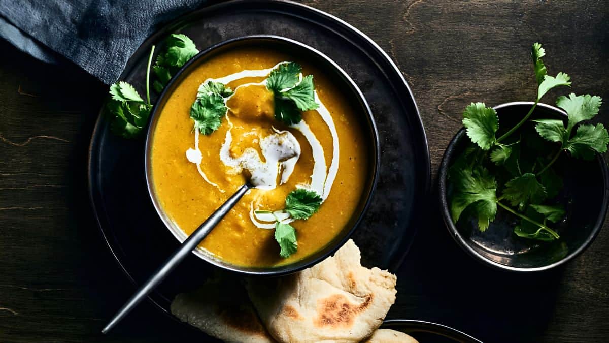 A bowl of creamy butternut squash curry soup garnished with a swirl of cream and fresh cilantro sits on a dark plate. Adjacent to the bowl is a piece of naan bread, and a sprig of cilantro rests beside the bowl on the plate. A dark napkin is partially visible in the background.
