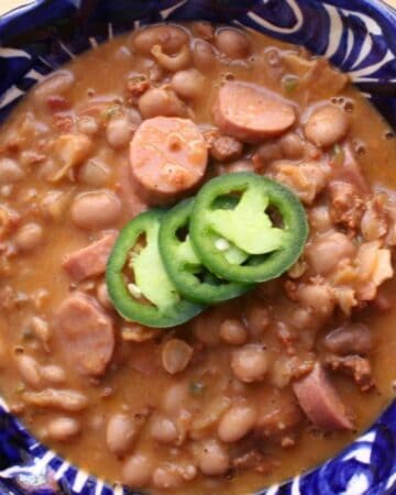 A blue and white ceramic bowl filled with a hearty Frijoles Charros stew topped with sliced jalapeños is placed on a wooden surface. Fresh cilantro is visible to the side, and a spoon rests nearby, making it one of the most irresistible bacon recipes for any meal.