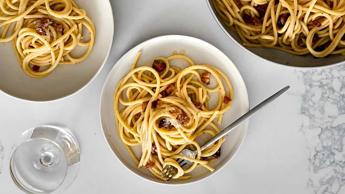 A flat lay of a table featuring three bowls of spaghetti carbonara. Two bowls are partially filled, and one is almost empty with a fork resting on the edge. A pot containing more spaghetti carbonara is also visible, along with a glass of water.