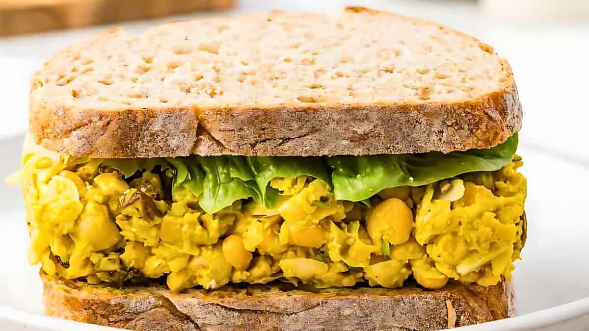 A sandwich made with two slices of whole grain bread, filled with a mixture containing chickpeas, spices, and green leafy vegetables, placed on a white plate. The filling appears vibrant and textured, contrasting with the earthy tone of the bread.