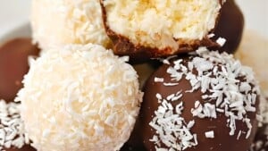 A close-up of assorted truffles coated in chocolate and shredded coconut. One of the truffles with shredded coconut coating is bitten, revealing a creamy inner filling. The truffles are stacked together, highlighting their rich and textured surfaces.