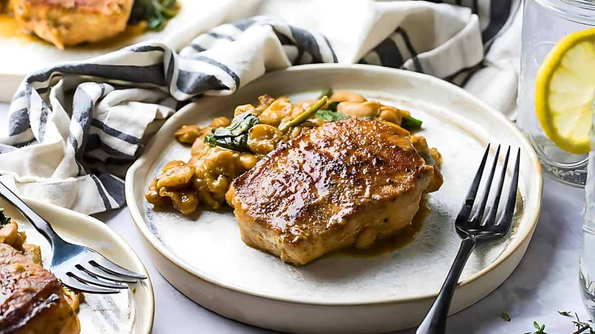 A meal featuring a seared piece of meat, likely pork, sits on a white plate beside a creamy bean and spinach mixture. A black fork lies on the plate's edge. The background includes a striped fabric napkin and a glass with a lemon slice.