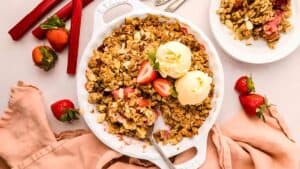 A white baking dish filled with strawberry rhubarb crisp topped with golden oat crumble and two scoops of vanilla ice cream, garnished with fresh strawberries. A smaller plate with a serving of the crisp and a fork is beside it. Scattered strawberries and a pink cloth surround the dishes.