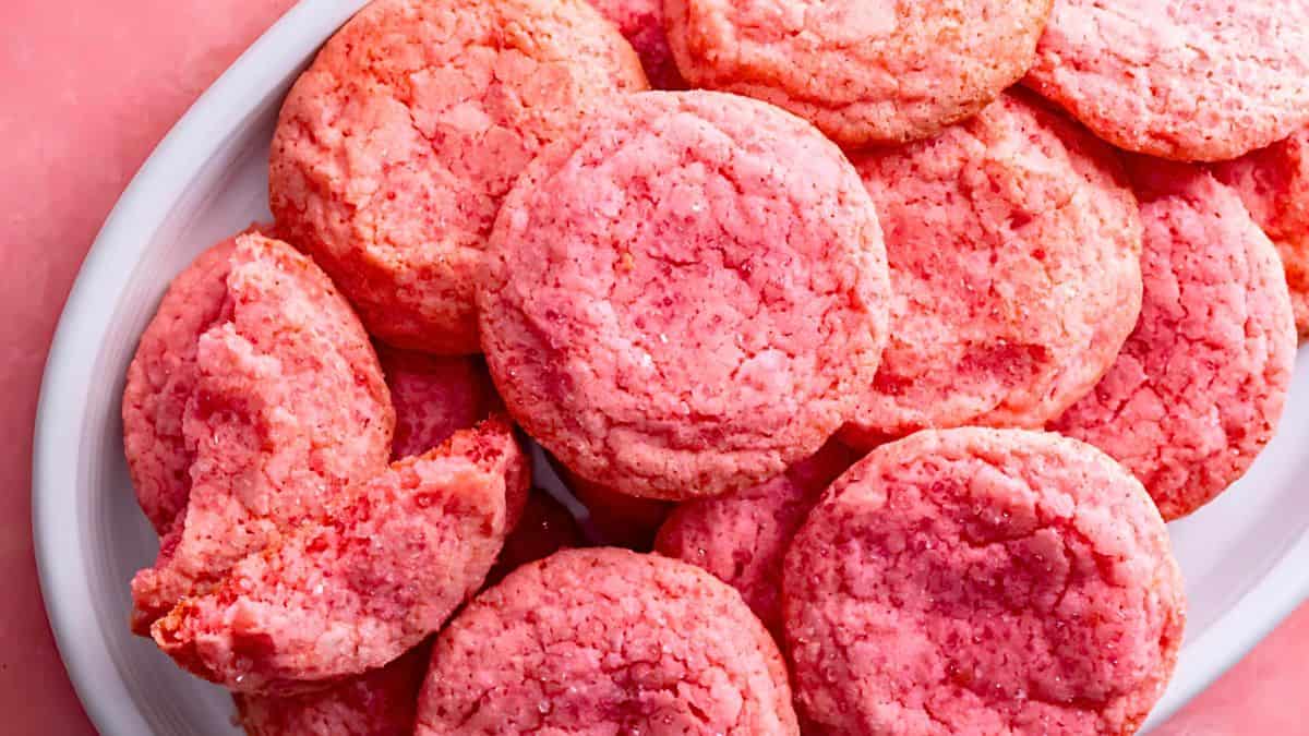 A white plate filled with vibrant pink cookies, some with a crumbly texture. One of the cookies at the edge of the plate appears broken in half, revealing a soft interior. The background is a matching pink surface, complementing the color of the cookies.