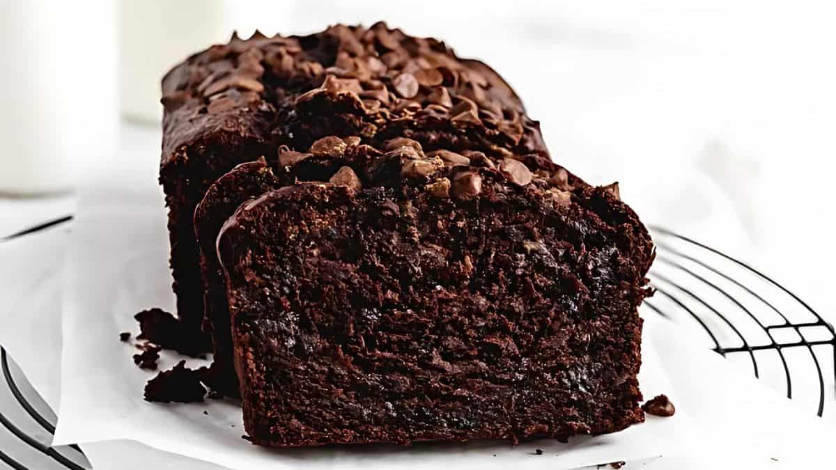 A loaf of moist, chocolate banana bread sits on a cooling rack, partially sliced to reveal its rich, dark, and dense texture. The bread is topped with chocolate chips, adding a layer of sweetness and richness to the visually appealing baked treat.