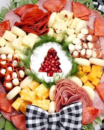 A holiday-themed charcuterie board arranged in a wreath shape with various meats, cheeses, and a small dish of dip topped with pomegranate seeds at the center. The board is decorated with checkered ribbons, greenery, and festive ornaments.