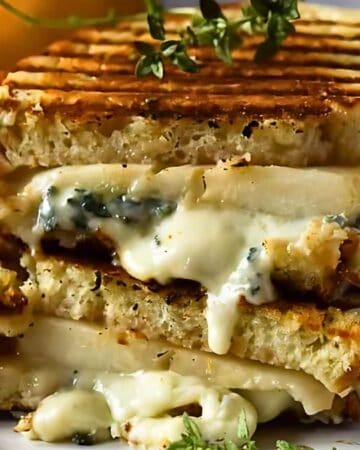 A close-up of a grilled cheese sandwich stacked in halves on a white plate. The sandwich is oozing with melted cheese and is garnished with a sprig of herbs. In the background, there is a blurred jar and some fruit.